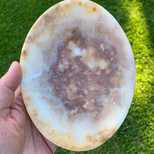 May include: An oval-shaped stone dish featuring a polished surface with a natural marbled pattern. The stone displays a blend of creamy white, translucent beige, and deep reddish-brown mineral inclusions throughout its smooth, rounded form.