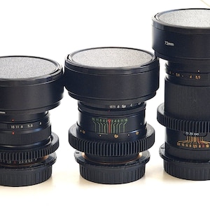 Puede incluir: Tres objetivos de cámara negros con tapas plateadas. Cada objetivo tiene diferentes tamaños y marcas, incluyendo "72mm". Los objetivos tienen varias escalas de medición, sobre un fondo blanco.