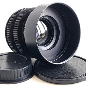 May include: A black Canon lens with a lens cap and a black lens hood. The lens has a silver ring with the text "NP-18" and "M.W." printed on it. The lens hood has the number "79597" printed on it.