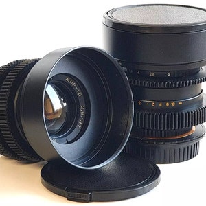 May include: A black camera lens with a 72mm filter attached. The lens has a black lens cap and a black lens hood. The lens has a depth of field scale and a focus ring. The lens is a Mir-1B 2.8/37.