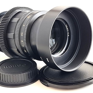 May include: A black Canon camera lens with a lens hood and two lens caps. The lens has a gear ring and markings for aperture settings from 2.8 to 22. The lens hood has the text "TAMP-IIIA" printed on it.