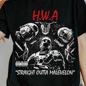 Op de afbeelding: Zwart T-shirt met een grafisch ontwerp met de tekst "H.W.A" in het rood boven een gedetailleerde illustratie van soldaten en de zin "STRAIGHT OUTTA MALEVELON". Het shirt heeft een ronde hals en korte mouwen.