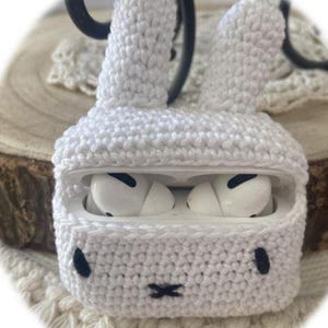 Peut inclure: Étui en crochet blanc en forme de lapin pour écouteurs sans fil. L'étui a un petit nez et des yeux noirs, et les écouteurs sont visibles à l'intérieur.