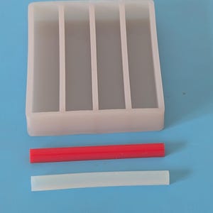 May include: A rectangular, translucent silicone mold with four vertical dividers. Two rectangular silicone sticks, one red and one white, are positioned below the mold. The items are on a blue surface.