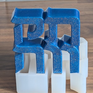 May include: A blue resin sculpture with glitter accents, possibly a logo or initials, is displayed in a white silicone mold. The sculpture has a glossy finish and is set against a wooden surface.