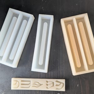 May include: A collection of silicone molds in various sizes and shapes, designed for crafting. The molds are white and beige, with a rectangular design and rounded interior channels. A small, rectangular piece with etched symbols is also present.
