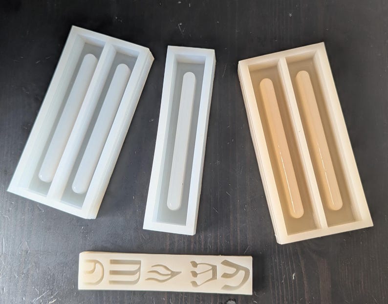 May include: A collection of silicone molds in various sizes and shapes, designed for crafting. The molds are white and beige, with a rectangular design and rounded interior channels. A small, rectangular piece with etched symbols is also present.