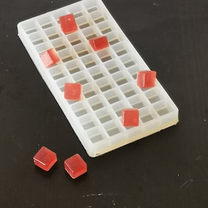 Puede incluir: Una bandeja de plástico transparente con una cuadrícula de compartimentos cuadrados. La bandeja está llena de pequeños caramelos de gelatina cuadrados rojos. Algunos de los caramelos están en la bandeja, y otros están esparcidos sobre una superficie negra.