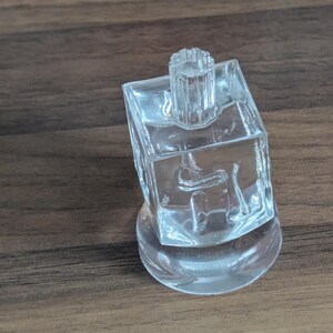 Puede incluir: Un objeto decorativo de vidrio transparente con una parte superior cuadrada y una base redonda. La parte superior tiene un peque&ntilde;o dise&ntilde;o cil&iacute;ndrico. El objeto est&aacute; sobre una superficie de madera.