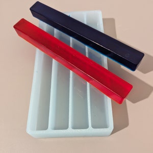 Puede incluir: Un molde de silicona blanco con cuatro ranuras rectangulares, dos de las cuales están llenas de resina. Una pieza de resina es roja y la otra es azul.