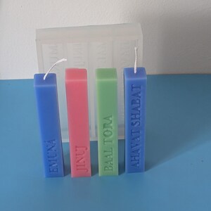 May include: Four colourful, rectangular candles with white wicks. The candles are blue, pink, green, and blue, each with a word embossed on the front: EMUNA, JINUJ, BAAL TORA, and AHAVAT SHABAT. A clear mould is in the background.