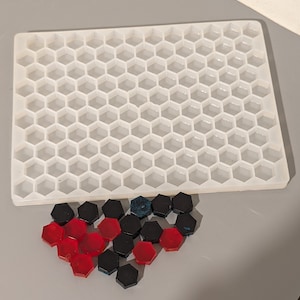 Può includere: Una griglia esagonale in plastica bianca con tessere esagonali rosse e nere sparse sotto. Le tessere hanno la forma di un cuore.