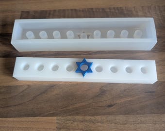 May include: Two white rectangular silicone molds for crafting a menorah. One mold has a Star of David in blue and the other has a Star of David in the center. The molds have circular indentations for candles.