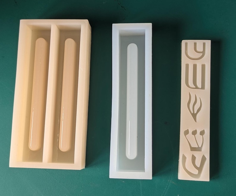 May include: Three silicone molds in a row. The first mold is rectangular with two rounded, cylindrical cavities. The second mold is a long, rectangular shape with a single, rounded cavity. The third mold is a rectangular shape with Hebrew letters and symbols.