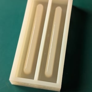 May include: A rectangular, off-white silicone mold with two long, rounded cavities. The mold is designed for creating bar-shaped items, possibly for crafts or soapmaking. The background is a solid teal color.