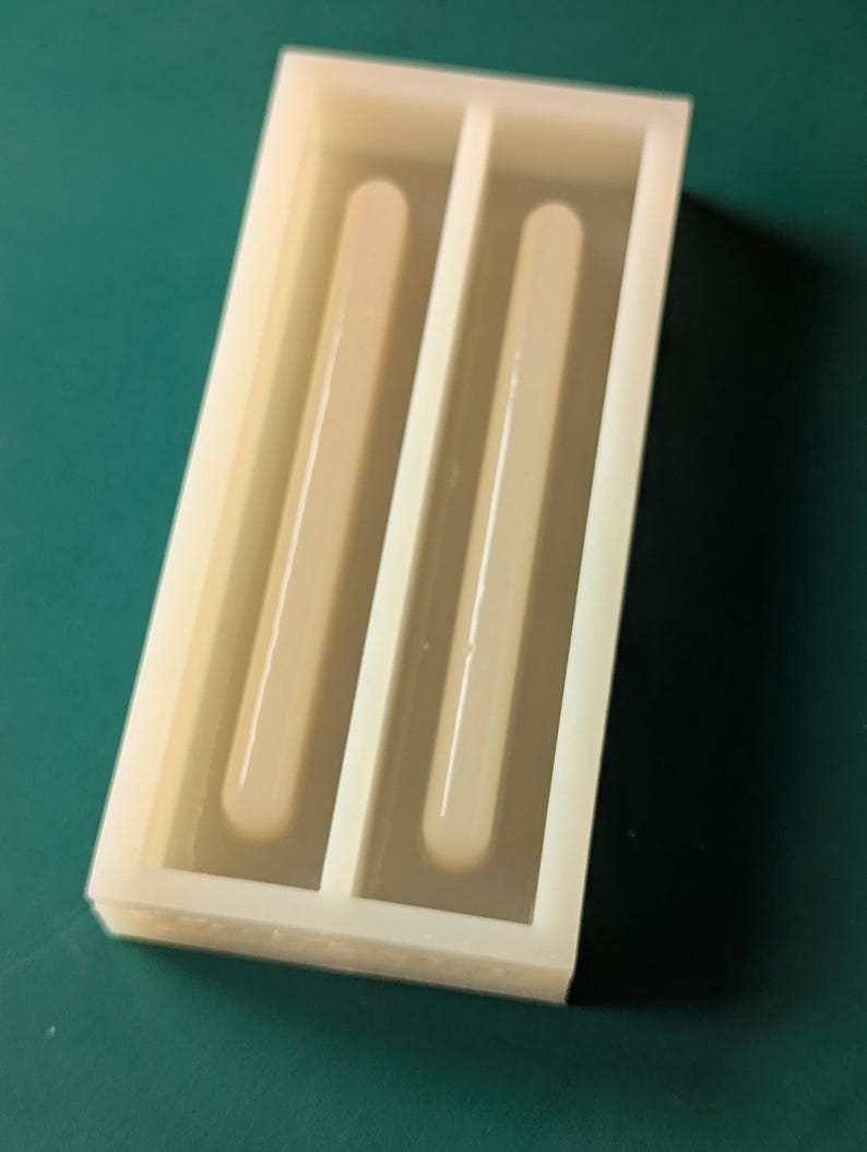 May include: A rectangular, off-white silicone mold with two long, rounded cavities. The mold is designed for creating bar-shaped items, possibly for crafts or soapmaking. The background is a solid teal color.
