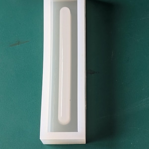May include: A white rectangular silicone mold with a long, rounded-end channel running down the center. The mold is on a green surface. The mold is likely for creating resin or other craft projects.