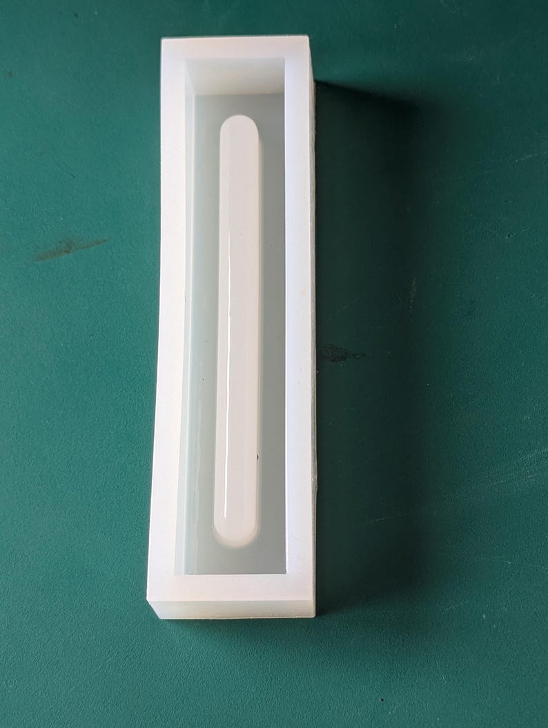 May include: A white rectangular silicone mold with a long, rounded-end channel running down the center. The mold is on a green surface. The mold is likely for creating resin or other craft projects.