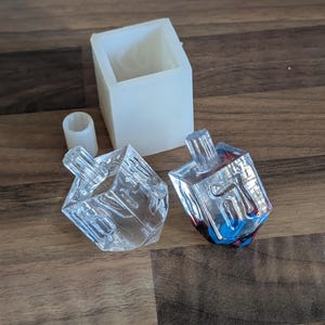 Peut inclure: Deux toupies dreidel transparentes à facettes, avec une base carrée et un dessus cylindrique transparent. Un moule carré blanc et un petit moule cylindrique blanc sont également présents. Les toupies ont un design géométrique clair.