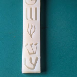 May include: A rectangular, off-white silicone mold with Hebrew letters. The mold features the letters arranged vertically. The background is a solid teal color.