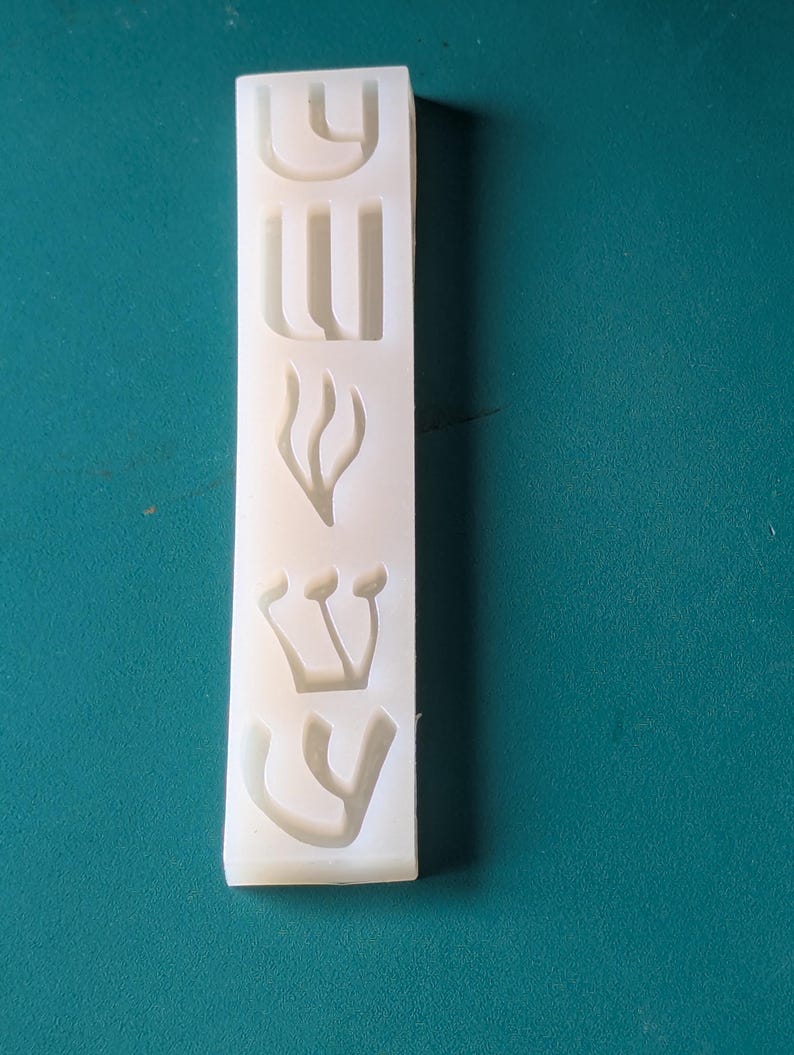 May include: A rectangular, off-white silicone mold with Hebrew letters. The mold features the letters arranged vertically. The background is a solid teal color.