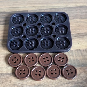 Op de afbeelding: Een siliconen mal met 12 ronde holtes voor het maken van knopen. Er zijn 10 bruine plastic knopen met elk vier gaten.