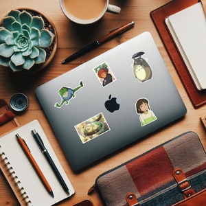 May include: A laptop computer with five stickers on it. The stickers are of a frog, a bowl of food, a girl with long black hair, a character from the movie "My Neighbor Totoro", and a character from the movie "Spirited Away".