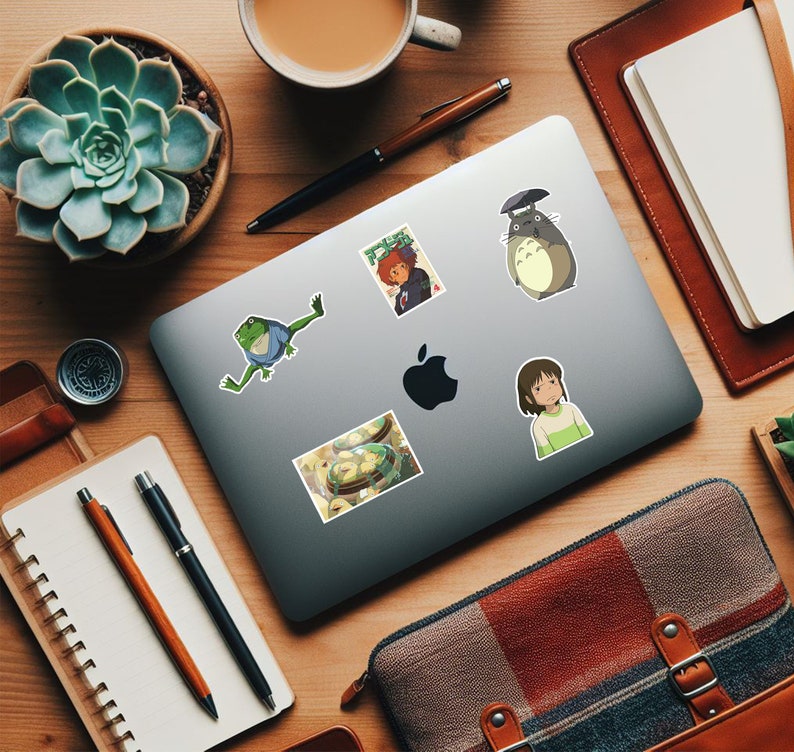 May include: A laptop computer with five stickers on it. The stickers are of a frog, a bowl of food, a girl with long black hair, a character from the movie "My Neighbor Totoro", and a character from the movie "Spirited Away".