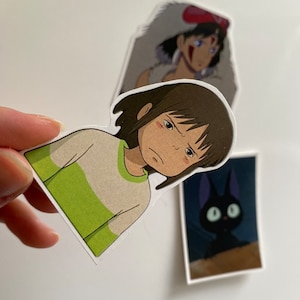 May include: Three stickers featuring characters from the animated film Spirited Away. The first sticker shows Chihiro, a young girl with brown hair, wearing a green and white shirt. The second sticker shows a black cat with large, yellow eyes. The third sticker shows a young woman with long, dark hair, wearing a red headband and a white fur coat.