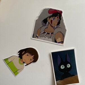 May include: Three stickers featuring characters from the animated film 'Princess Mononoke'. The first sticker shows a young woman with brown hair and a white wolf fur cloak. The second sticker shows a young girl with brown hair wearing a green and white shirt. The third sticker shows a black cat with large green eyes.