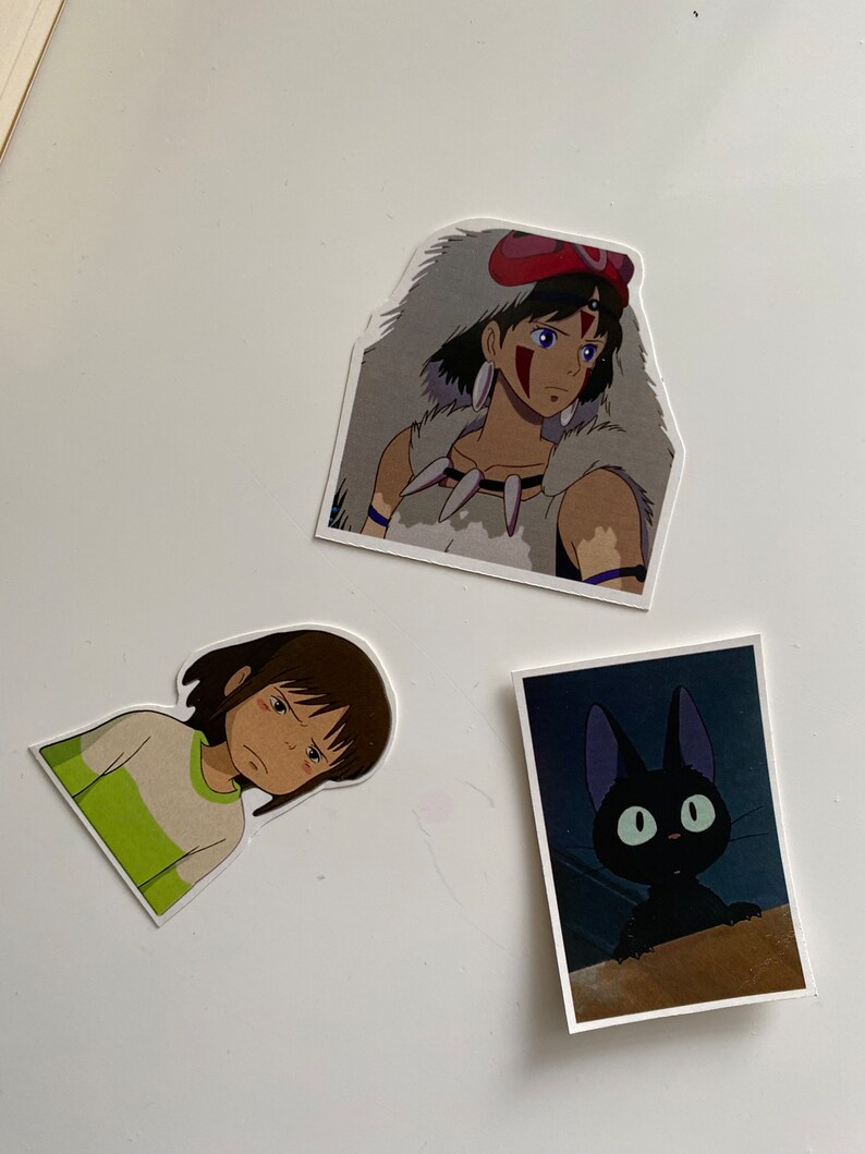 May include: Three stickers featuring characters from the animated film 'Princess Mononoke'. The first sticker shows a young woman with brown hair and a white wolf fur cloak. The second sticker shows a young girl with brown hair wearing a green and white shirt. The third sticker shows a black cat with large green eyes.