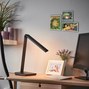 May include: A black desk lamp with a flexible arm is on a wooden desk with a white framed photo, a keyboard, and a computer monitor. There are three framed posters on the wall behind the desk, featuring characters from the animated film "My Neighbor Totoro".