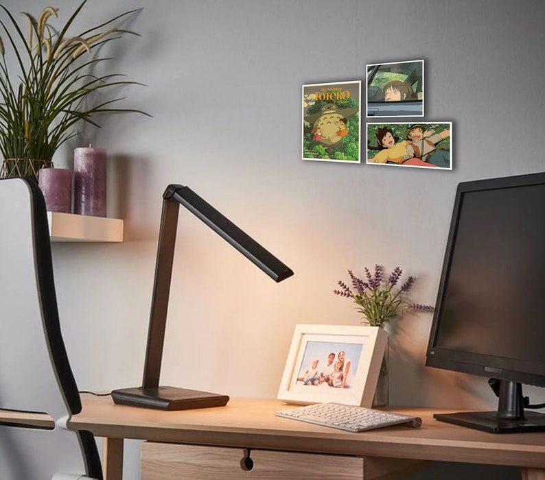 May include: A black desk lamp with a flexible arm is on a wooden desk with a white framed photo, a keyboard, and a computer monitor. There are three framed posters on the wall behind the desk, featuring characters from the animated film "My Neighbor Totoro".