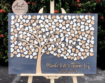Può includere: Un libro degli ospiti a forma di albero di cuori in legno su sfondo grigio. L'albero ha un tronco e rami marroni con cuori di legno bianchi e marroni. Il testo "Minh Loi & Thien Ly" e "21.01.2024" è scritto sotto l'albero.
