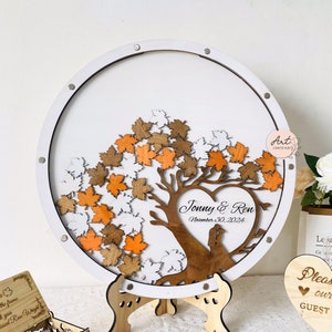 May include: A white circular wooden guest book with a brown tree and heart design. The tree has orange and white leaves. The heart has the names "Jonny & Ren" and the date "November 30, 2024".