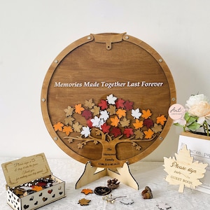 May include: A wooden guest book alternative in the shape of a tree with fall-colored leaves. The leaves are attached with magnets and can be signed by guests. The sign reads "Memories Made Together Last Forever".
