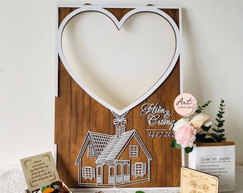 Può includere: Una cornice di legno a forma di cuore con un cuore bianco ritagliato al centro. La cornice è decorata con una piccola casa e il testo "Hiền & Cương 14.07.202" in bianco. La cornice è su una superficie bianca con una piccola scatola di coriandoli colorati e alcuni oggetti decorativi.