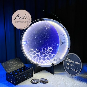 May include: An illuminated moon and star-shaped wedding sign with the names "Marry & John" and the date "07.30.2025". The sign is supported by a black stand and surrounded by other wedding decor.