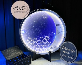 May include: A decorative illuminated moon and star-shaped wedding sign with the names "Marry & John" and the date "07.30.2025". The sign is supported by a black stand and surrounded by other wedding decor.