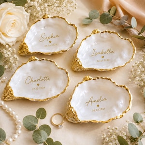 May include: Four white oyster shell trinket dishes with gold-painted edges. Each shell features a custom name in gold script, a small heart icon, and a date: Sophia 05.16.2026, Isabella 06.21.2026, Charlotte 07.10.2026, and Amelia 08.03.2026.