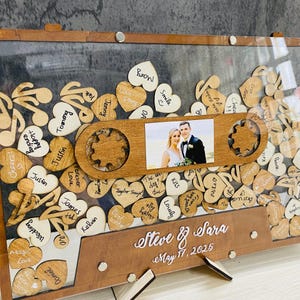 May include: A wooden wedding guest book with a clear acrylic cover. The guest book is shaped like a cassette tape and has a small photo frame in the centre. The photo frame shows a bride and groom. The guest book is decorated with wooden hearts and music notes. The hearts and music notes have handwritten messages on them. The guest book is signed "Steve & Sara" and "May 17, 2025".