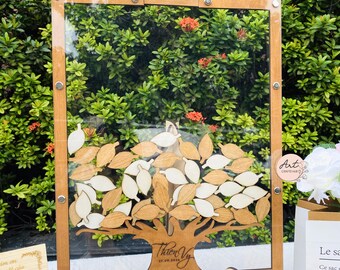 Può includere: Un albero di legno per libro degli ospiti con una cornice in acrilico trasparente. L'albero ha un tronco e rami marroni con foglie bianche e marroni. Le foglie sono progettate per essere firmate dagli ospiti. La cornice è montata su un cavalletto di legno. Il testo "Thien Vy 27.09.2020" è inciso sul tronco dell'albero.