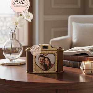 May include: A brown and gold camera-shaped photo frame with a heart-shaped photo of a couple. The frame sits on a dark wooden table, with a vase of white flowers and a lit candle nearby. The text "Art Crafts Hub" is visible.