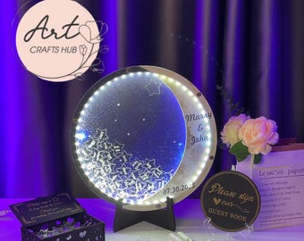 May include: A round, illuminated wedding guest book with a crescent moon design. The moon is filled with a starry night scene and the names "Marry & John" with the date "07.30.2025". Includes a guest book sign and a small box.