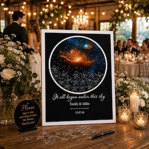 May include: A white-framed guest book shadow box featuring a circular space nebula print, filled with small star-shaped tokens. The text reads: It all began under this sky, Emily & Mike, 12.07.26. A small black sign with gold text instructs guests to sign a star.