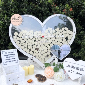 Puede incluir: Un libro de visitas de madera en forma de corazón blanco con un frente de acrílico transparente. El libro de visitas está lleno de pequeños corazones de madera. Una foto de una pareja se muestra en la esquina inferior derecha. El texto "TRONG TAN KHÁNH VÂN 20.04.2024" está escrito en la parte delantera del libro de visitas.