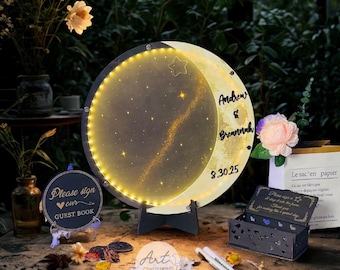 May include: A personalized moon-shaped light with a starry design and the names "Andrew & Breannah" and the date "9.30.25". The light is surrounded by a string of small lights. A guest book sign and a decorative box are also present.