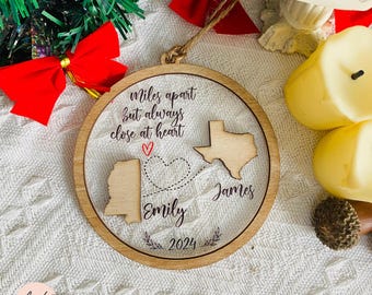 Puede incluir: Un adorno navideño de madera con fondo transparente. El adorno presenta dos estados, Texas y Alabama, conectados por una línea punteada con un corazón. El texto "miles apart but always close at heart" está escrito sobre los estados. Los nombres "Emily" y "James" están escritos debajo de los estados, y el año "2024" está escrito debajo de los nombres.