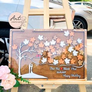 May include: A wooden guest book with a tree and leaves design. The leaves are in shades of brown, orange, and white. The text "Thu Hà ♥ Minh Phùi 28.04.2024 Happy Wedding" is written on the bottom of the guest book.