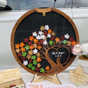 May include: A wooden guest book alternative in the shape of a tree with a heart at the base. The tree is decorated with fall-colored leaves and the heart has the text "Lily & Jessica 11-24-24" inscribed on it.
